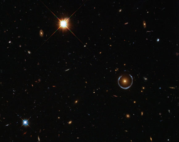 This Hubble image shows the Cosmic Horseshoe gravitational lens (to the right from the center): the newly-discovered ultramassive blackhole lies at the center of the orange galaxy; far behind it is a blue galaxy that is being warped into the horseshoe shaped ring by distortions in spacetime created by the immense mass of the foreground orange galaxy. Image credit: NASA / ESA / Hubble.