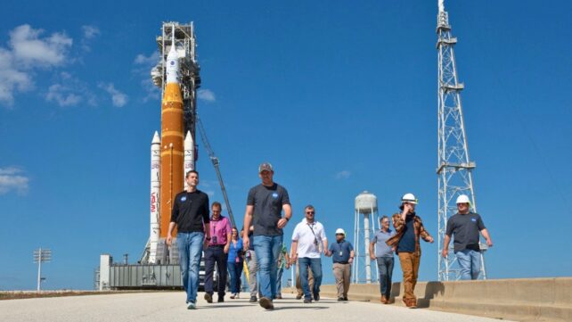 A group of people walking towards the viewer on a sunny day, with a rocket shown in high contrast in the background