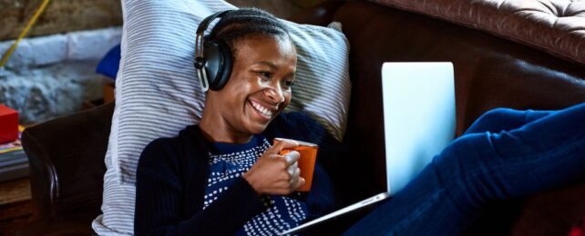 Young woman wearing headphones on laptop video call, drinking a coffee 
