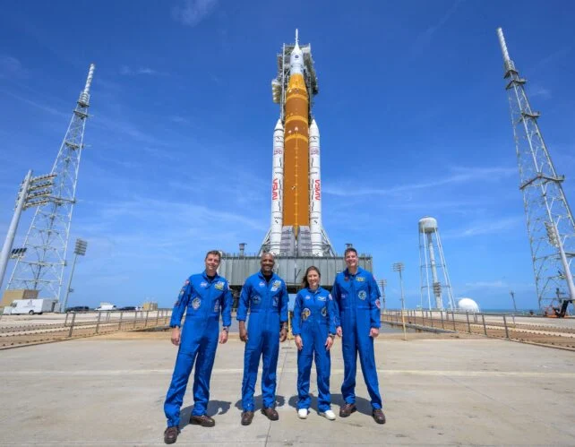 Four people in blue jumpsuits standing in front of rocket on conreted launch pad.