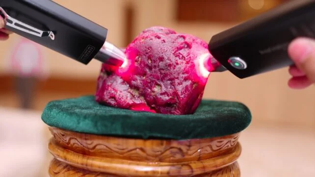 Two handheld lights shine through a huge rough ruby displayed on a green velvet stand.